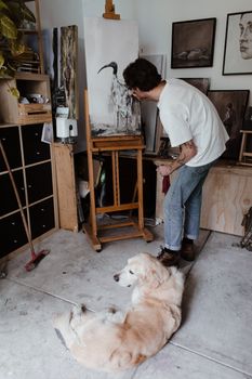 一位年轻男性艺术家穿着休闲服装，站在灯光工作室的画架旁绘画，旁边有一只可爱的金毛猎犬，他的侧面图看起来面目全非