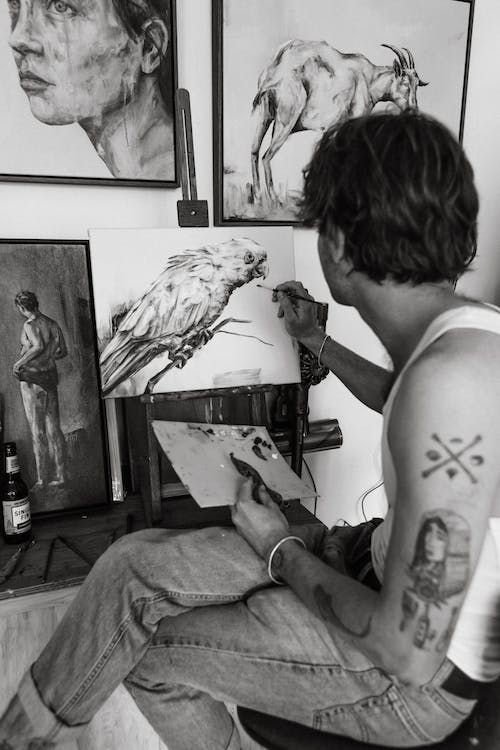 Side view of black and white young tattooed male sitting on chair and drawing various pictures with paintbrush in creative studio