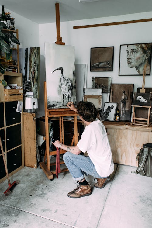 Side view of unrecognizable focused young male painter in stylish clothes sitting on haunches while drawing on easel in creative art studio