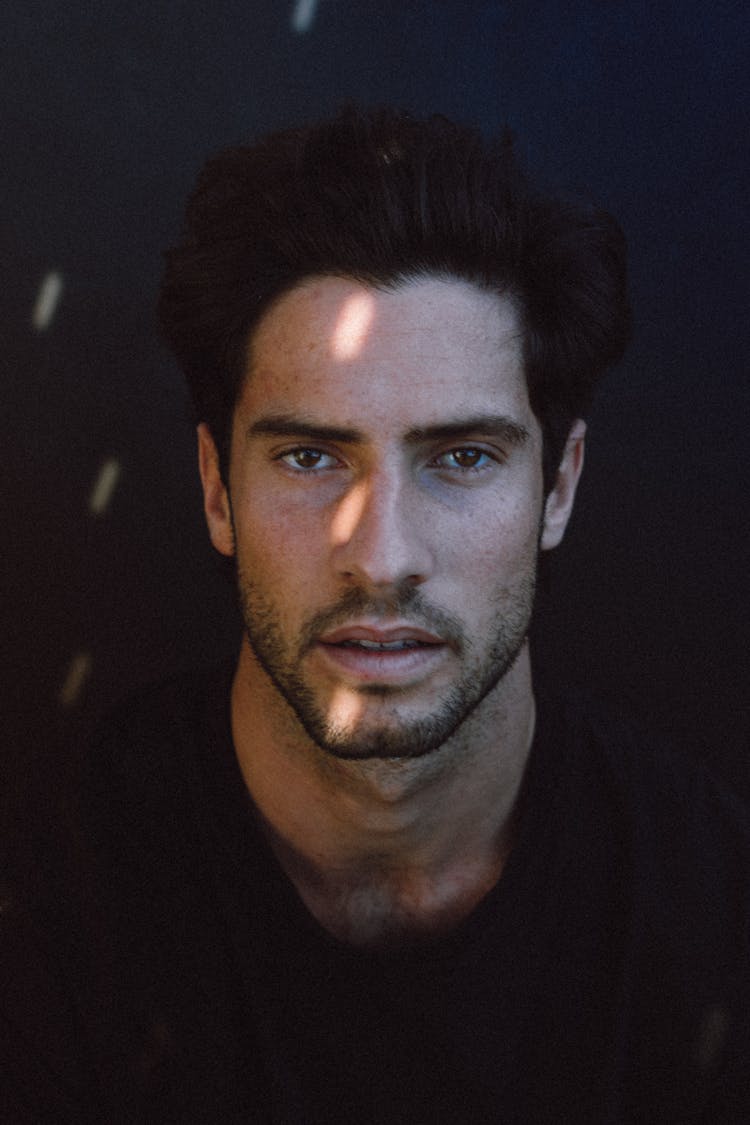 Contemplative Young Ethnic Man With Sunlight On Face Looking At Camera In Studio