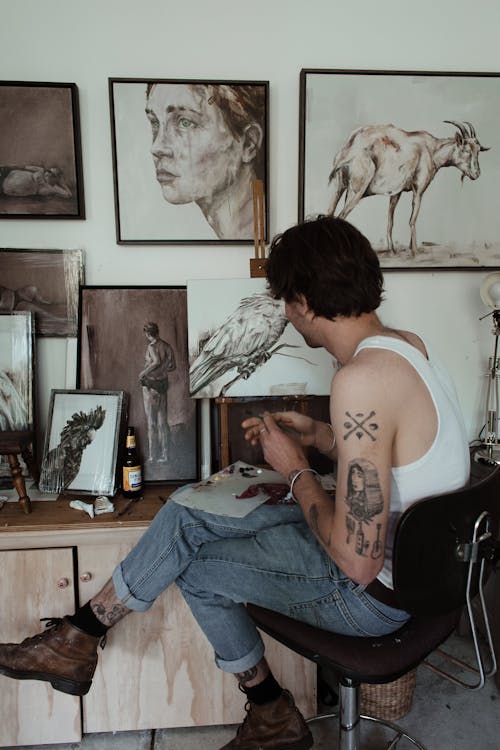 Side view of unrecognizable trendy tattooed male painter in casual outfit sitting on chair and mixing inks while drawing in creative workshop