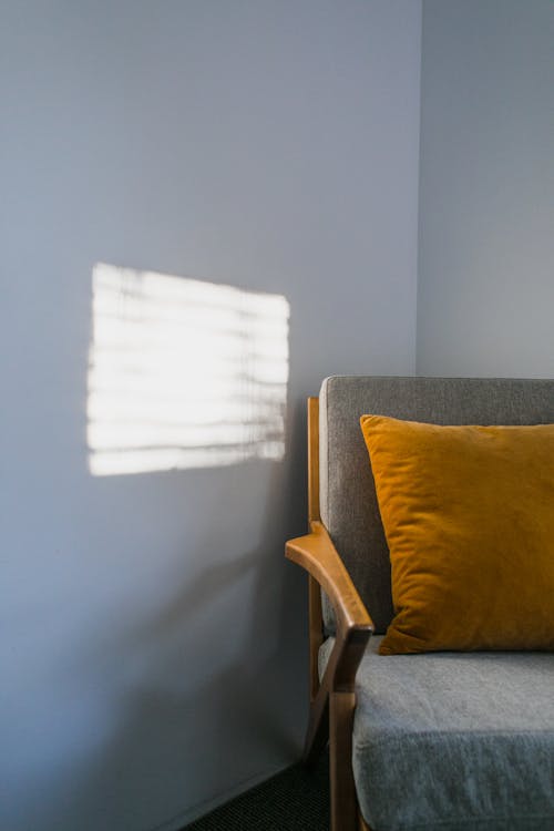 Comfortable gray armchair with orange pillow placed in corner of light room with bright sunlight on white wall at home