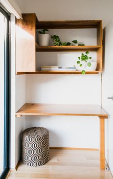 Empty table with pouf placed at white wall with wooden shelves with potted green flowers in light room at home