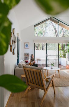 远处，一位女性侧坐在沙发上，现代宽敞的客厅里配有扶手椅和侧桌，家中点缀着绿叶