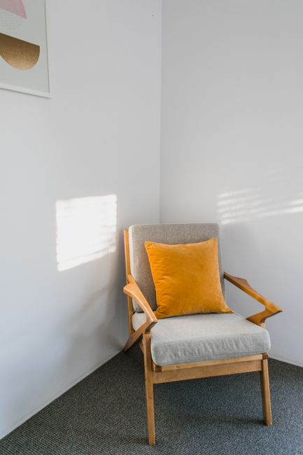 Comfortable wooden gray armchair with orange pillow placed near white wall with picture in light room with sunlight at home