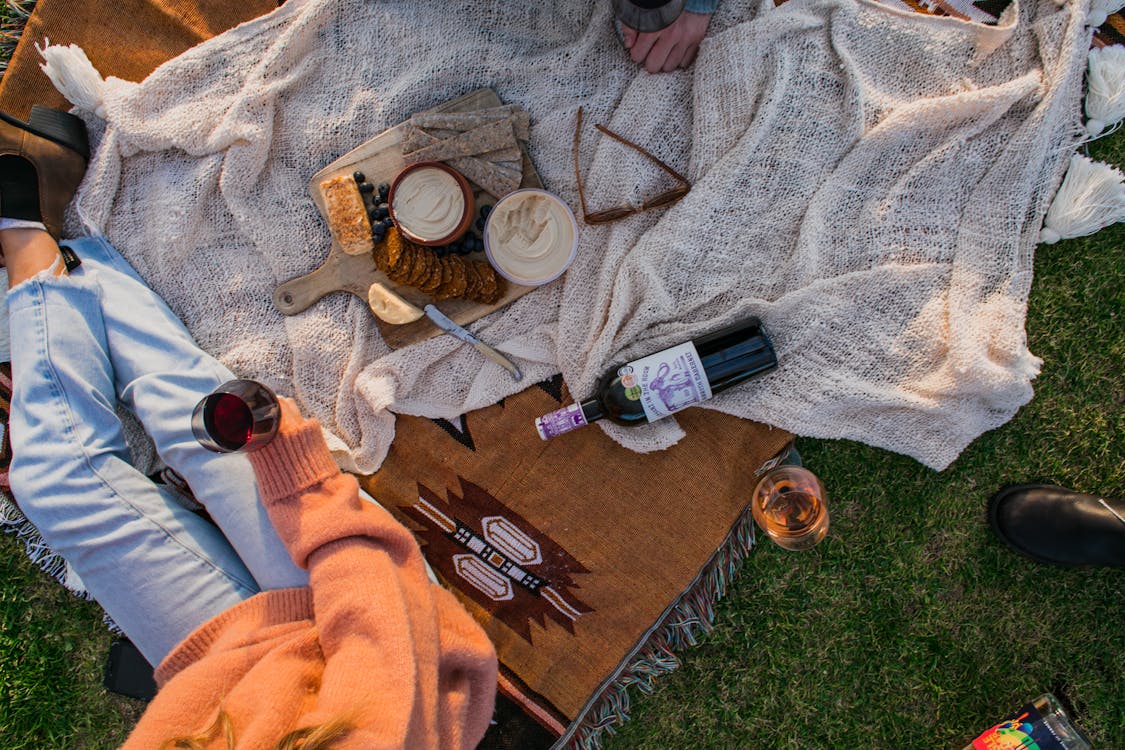 Free Top view of anonymous female with glass of wine sitting on plaid with food while having picnic on grassy lawn Stock Photo