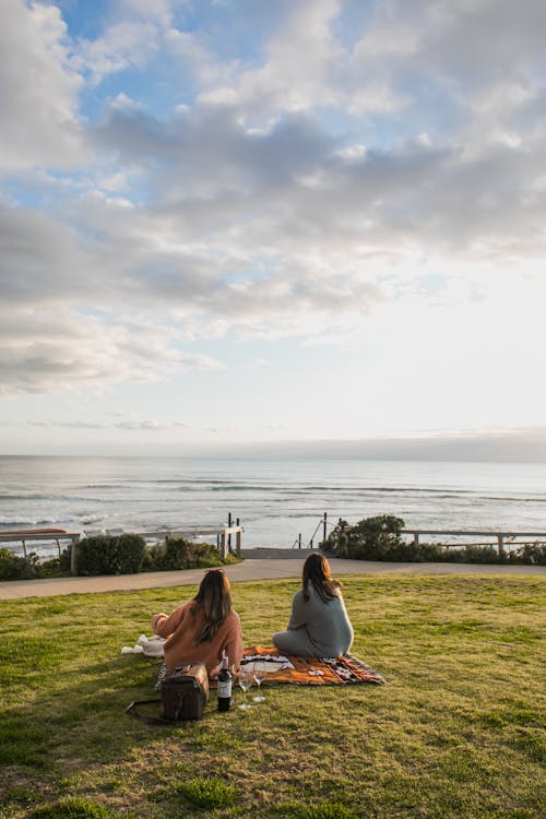 Free Back view of unrecognizable female friends sitting on grassy lawn near rippling water while having picnic with wine on waterfront Stock Photo