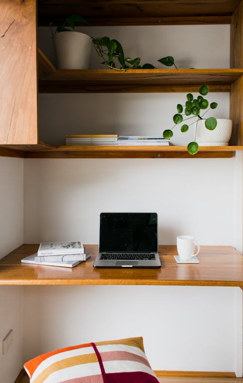 Modern netbook with black screen on wooden table with cup of hot drink near shelves with potted flowers at home