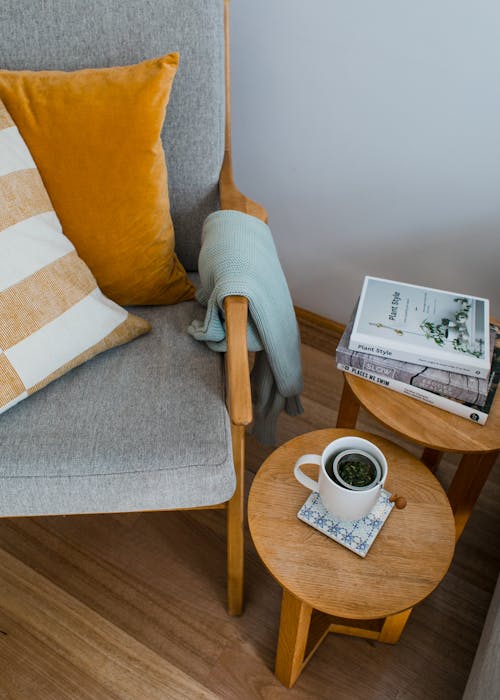 From above of comfortable armchair with colorful pillows placed near wooden side tables with books and cup of tea in living room