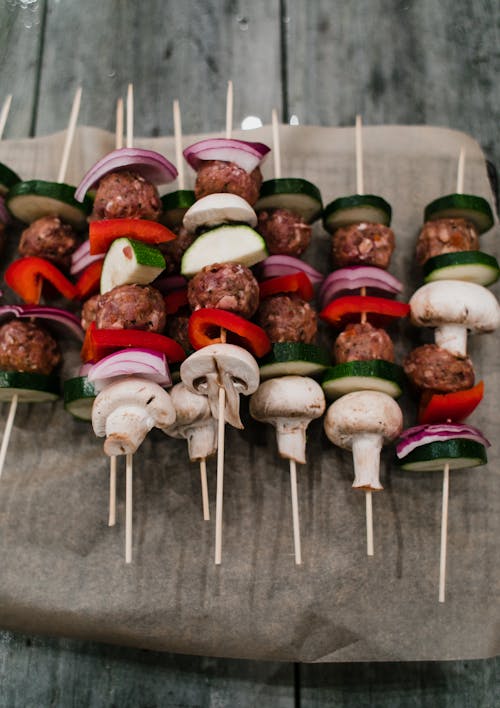 Free From above of tasty raw meatballs with mushrooms and red pepper with onion and zucchini on skewers on paper on wooden surface Stock Photo