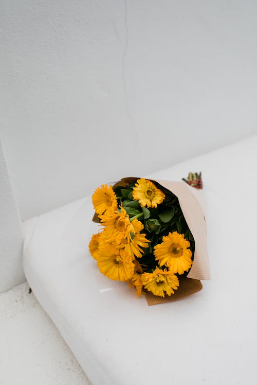 High angle of blossoming bouquet with yellow flowers wrapped in paper placed on white mattress in bright room near wall
