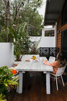 一位长发女子在摆满白色家具和盆栽植物的木制门廊上吃早餐，她若有所思地将目光移向别处