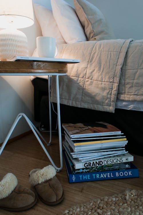 Stack of books placed on wooden floor near comfortable light bed and coffee table with lamp and white cup and soft slippers near wicker carpet