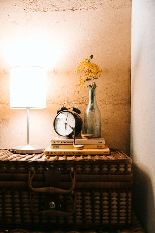 Creative rustic table made of vintage wicker suitcase with luminous lamp alarm clock and vase in dark bedroom