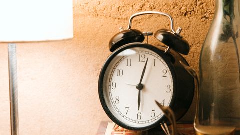 Old fashioned alarm clock placed on stacked books near luminous lamp on bedside table