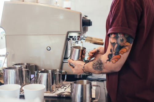 Crop anonymous male with tattoos preparing tasty aromatic coffee with coffee machine and pitcher
