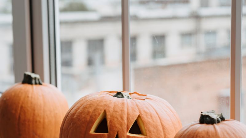 Three pumpkins including a carved jack-o'-lantern by a window, perfect for Halloween decor.