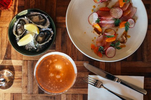 Free Top view glass of cold cocktail with bowl of fresh oysters with ice and lemon served on wooden table near plate of sliced fish garnished with herbs and radish Stock Photo