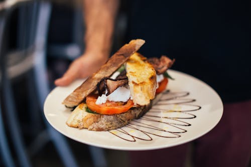 Free From above of crop unrecognizable waiter holding plate of appetizing sandwich with bacon tomatoes and poached eggs in crispy toasted bread Stock Photo