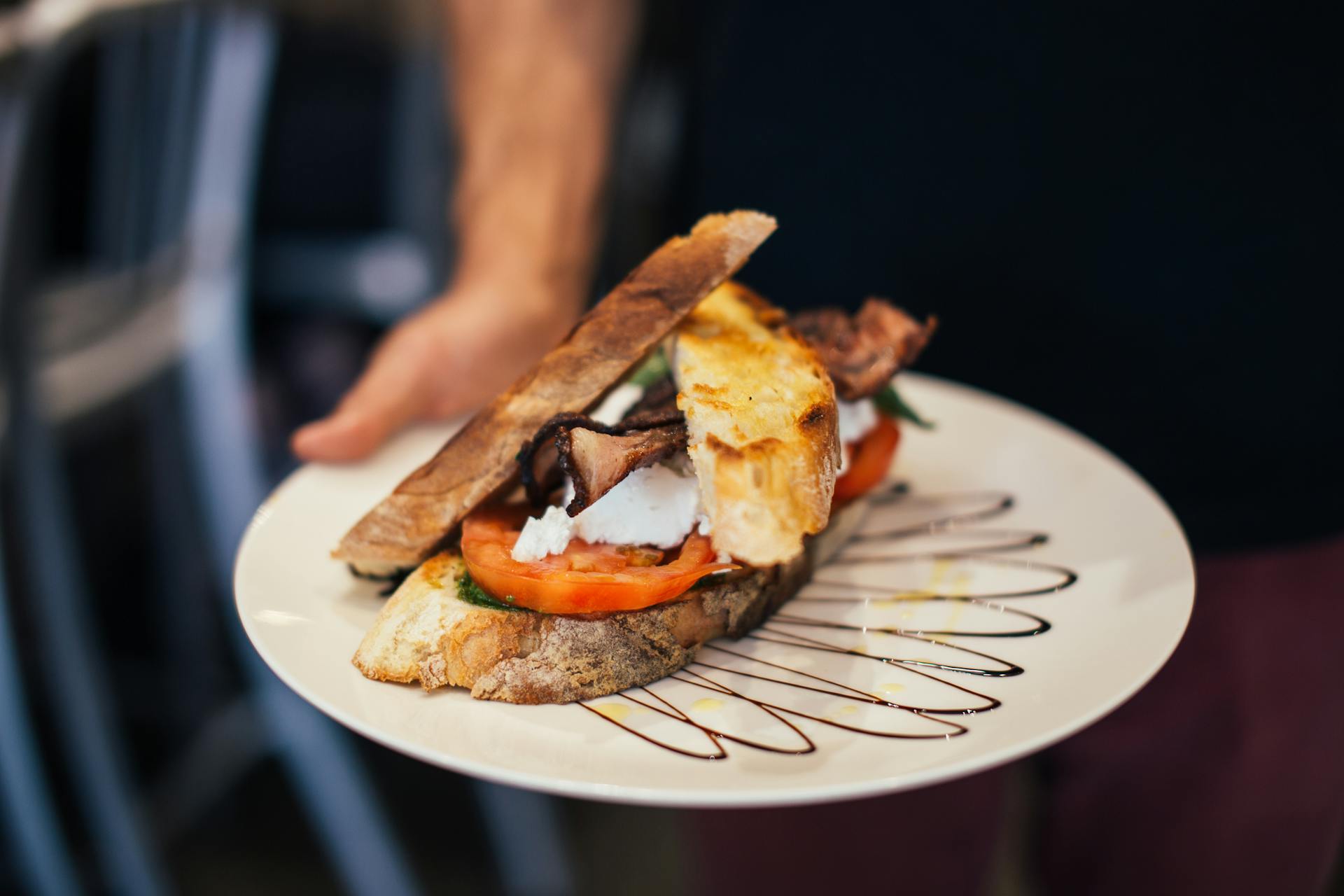 From above of crop unrecognizable waiter holding plate of appetizing sandwich with bacon tomatoes and poached eggs in crispy toasted bread