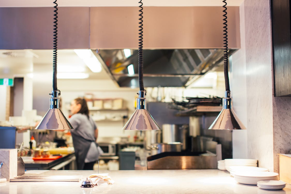 Side view of unrecognizable female chef in restaurant kitchen working behind counter with infrared lamps and serving kitchenware