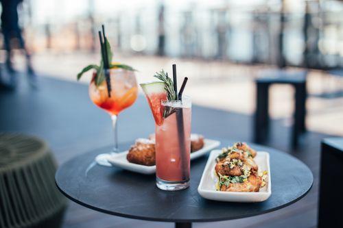 Free Glasses with colorful cocktails garnished with green herbs and straws with watermelon slice near white plates with delicious appetizers on round table in street restaurant in daytime Stock Photo