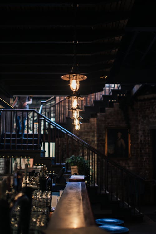 Glowing lamp hanging on ceiling above counter of bar in modern cafeteria with wooden stairway with railings and dim light