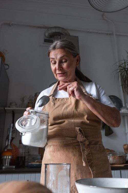Free Experienced senior woman baking in the kitchen, wearing an apron and handling flour. Stock Photo