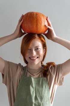Cheerful woman with red hair balances pumpkin on her head, showcasing a playful fall vibe.
