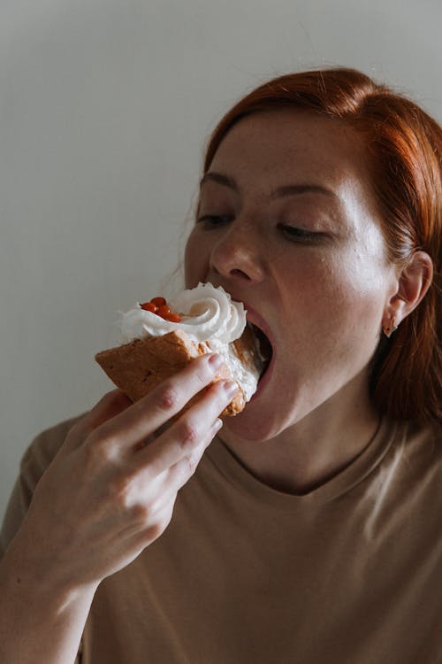 red-haired woman eats a large slice of cake covered in whipped cream with her hands.