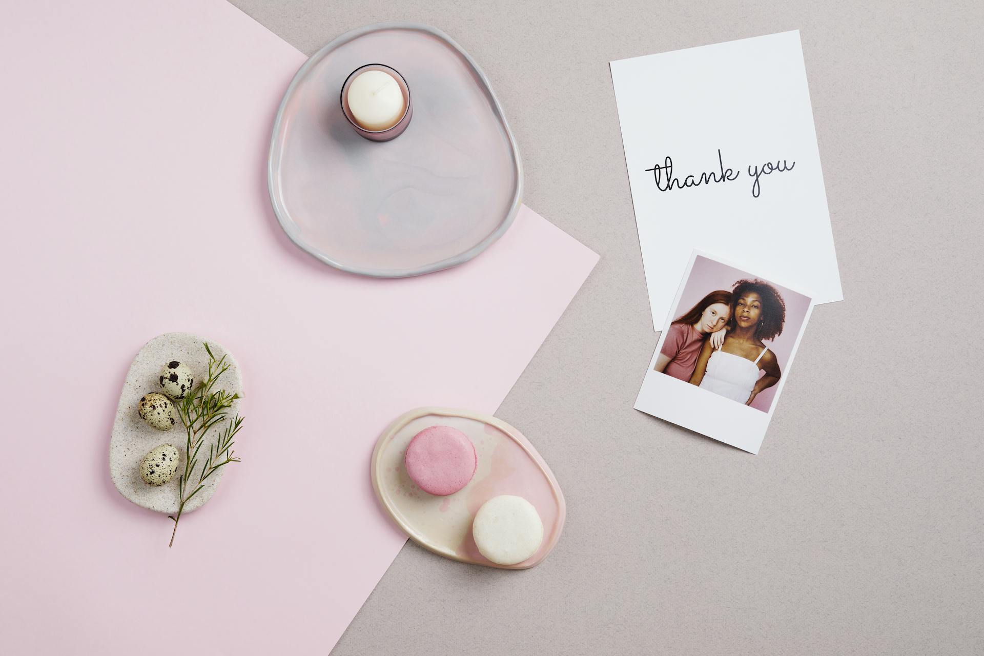 A minimalist flat lay with pink and white macarons, thank you card, and Polaroid photo on a pastel background.