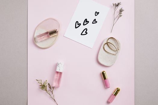 Elegant flat lay of perfume, lip gloss, and card with hearts on a pink background.