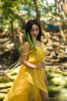 白天，一位迷人的年轻苗条少数族裔女性在森林中行走，她身穿飞扬的黄色背心裙，手持树枝和树叶，望向镜头