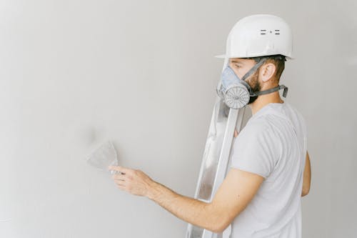 Man in White T-shirt Wearing White Gas Mask Using Flat Metal Spatula on the Wall