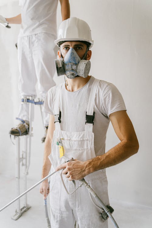 Man in White Crew Neck T-shirt Wearing Gas Mask and Hard Hat while Seriously Looking at Camera