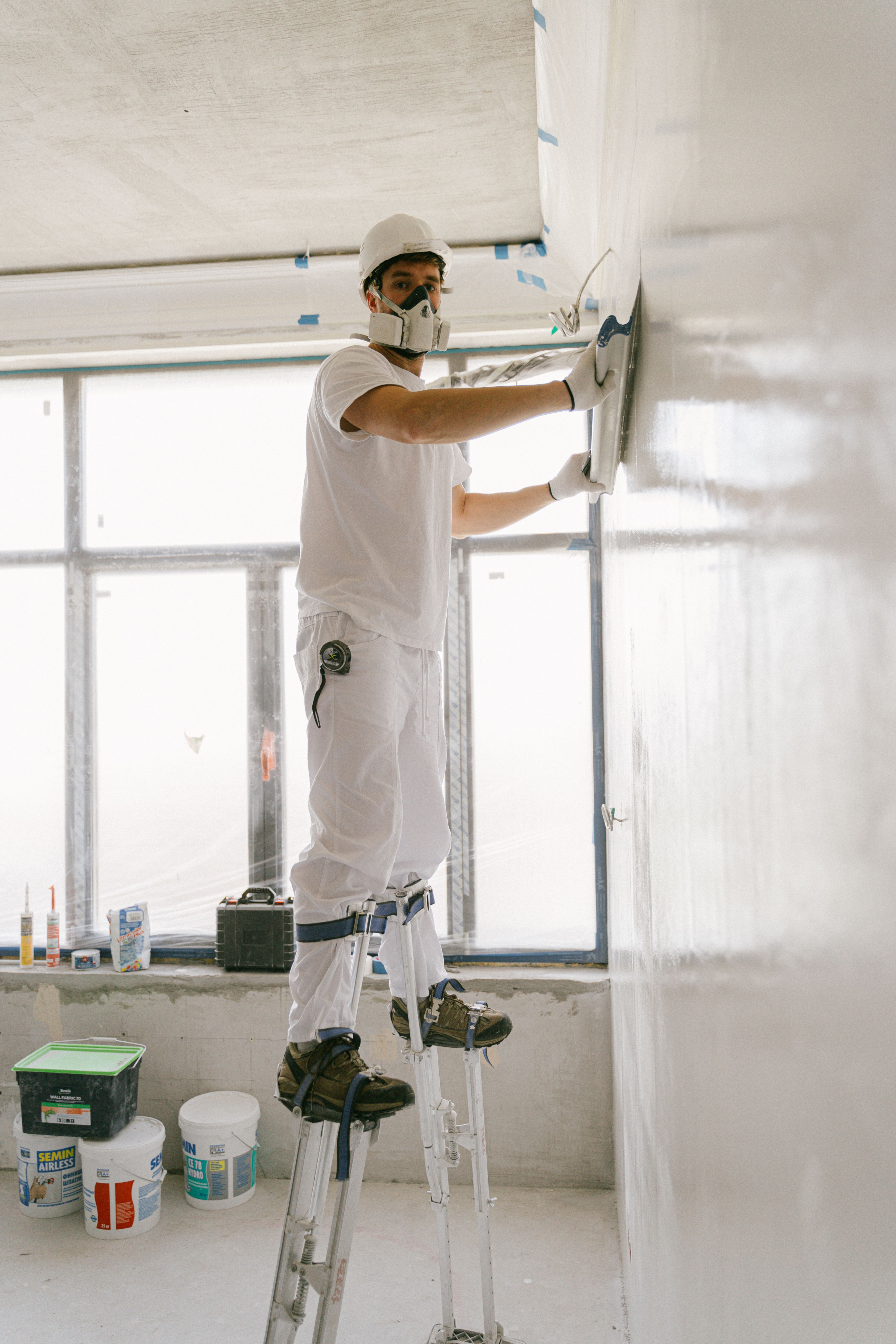 Worker wearing respiratory and head protection smooths a wall in a room under renovation