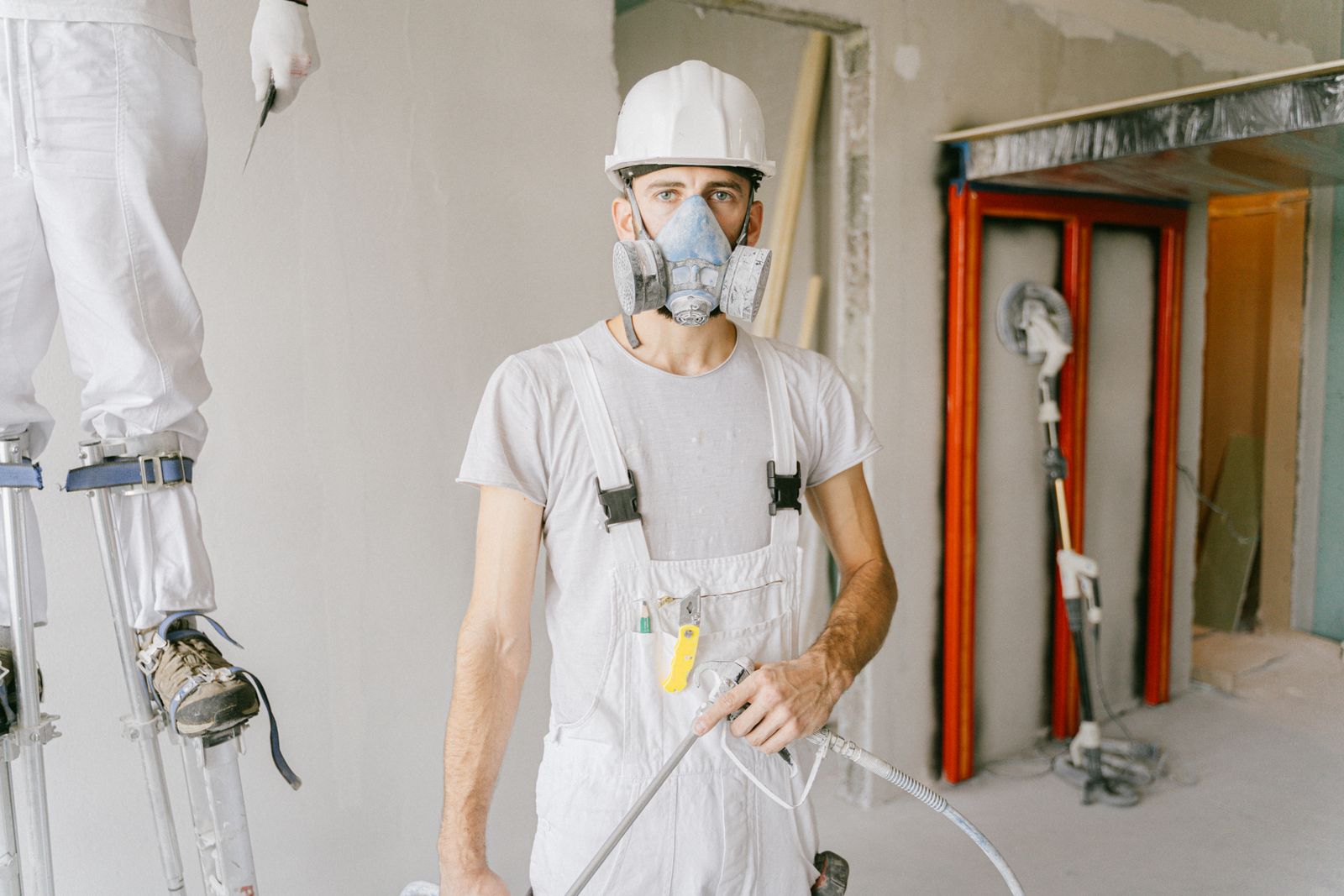 Technician in a hard hat and respirator holding spray equipment inside a gutted room under renovation