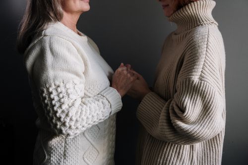 Free Two women warmly holding hands, showcasing textured knit sweaters indoors. Stock Photo
