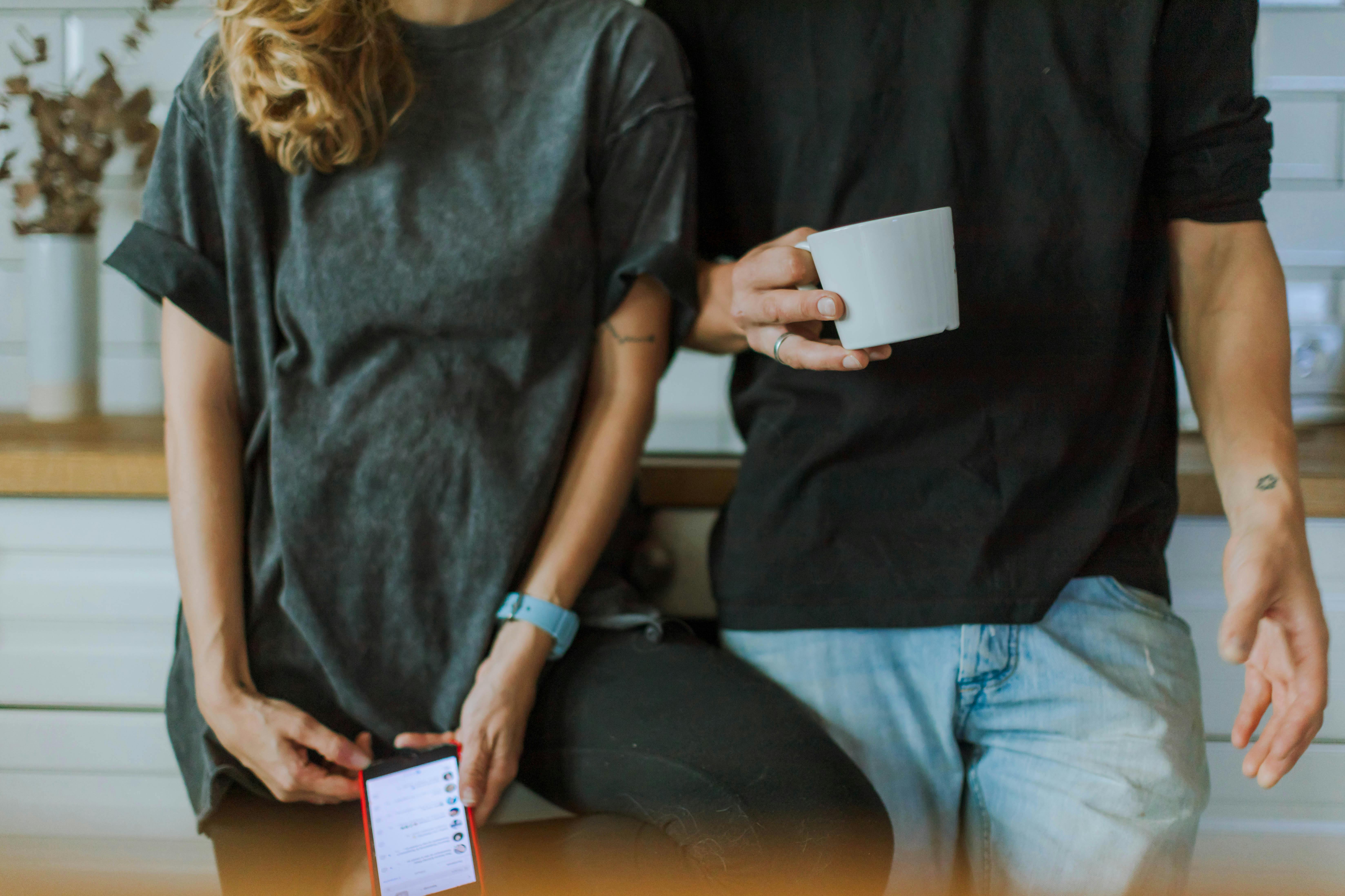 一对情侣在厨房里随意地聊着天，一个拿着智能手机，另一个端着咖啡杯