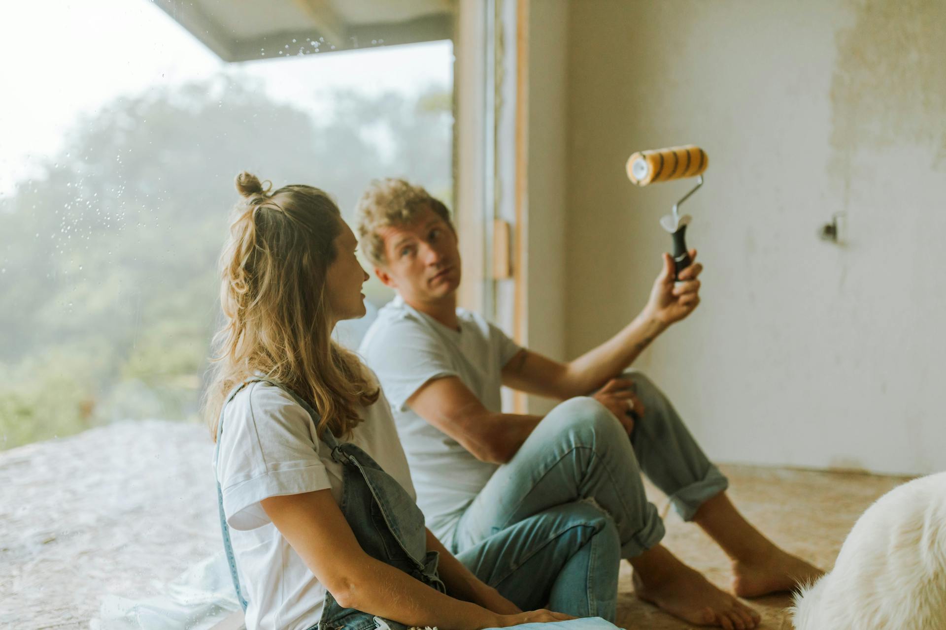 A young couple sits indoors planning their home renovation with a paint roller.