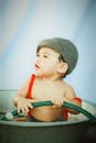 Adorable little boy in knitted hat sitting in metal basin with water and looking away