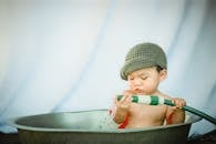 Charming baby boy looking closely at nozzle of garden hose while taking bath in old metal basin
