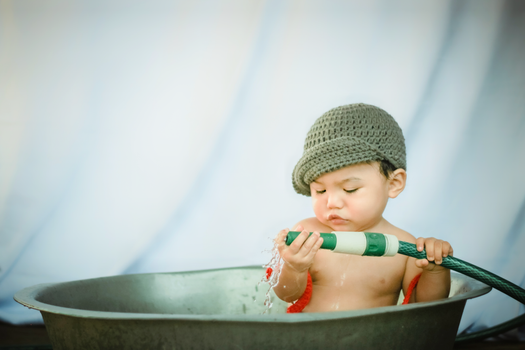 Charming baby boy looking closely at nozzle of garden hose while taking bath in old metal basin
