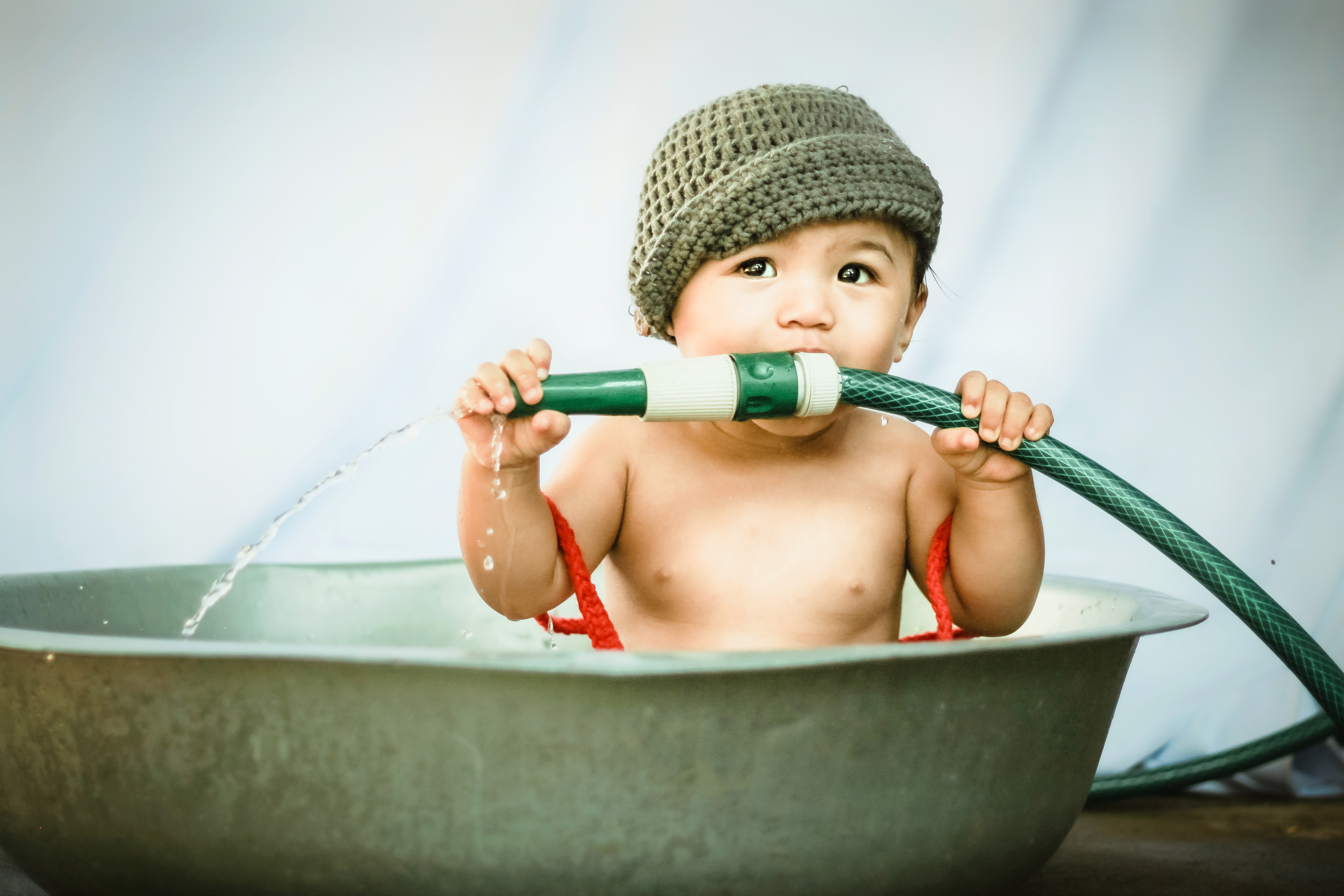 Attractive toddler sitting in basin and biting nozzle of hose while looking away