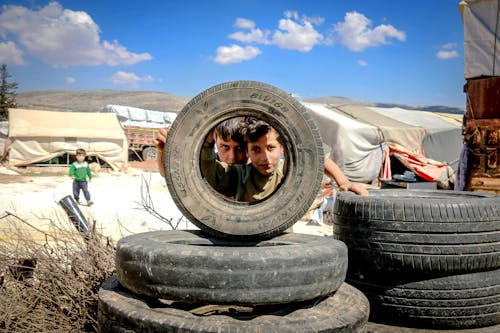 Pensive boys looking through big black wheel of vehicle under vibrant blue sky with clouds in encampment