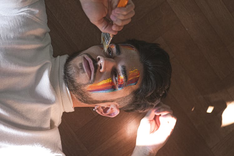 Man Lying On Floor And Painting Multicolored Lines On Face