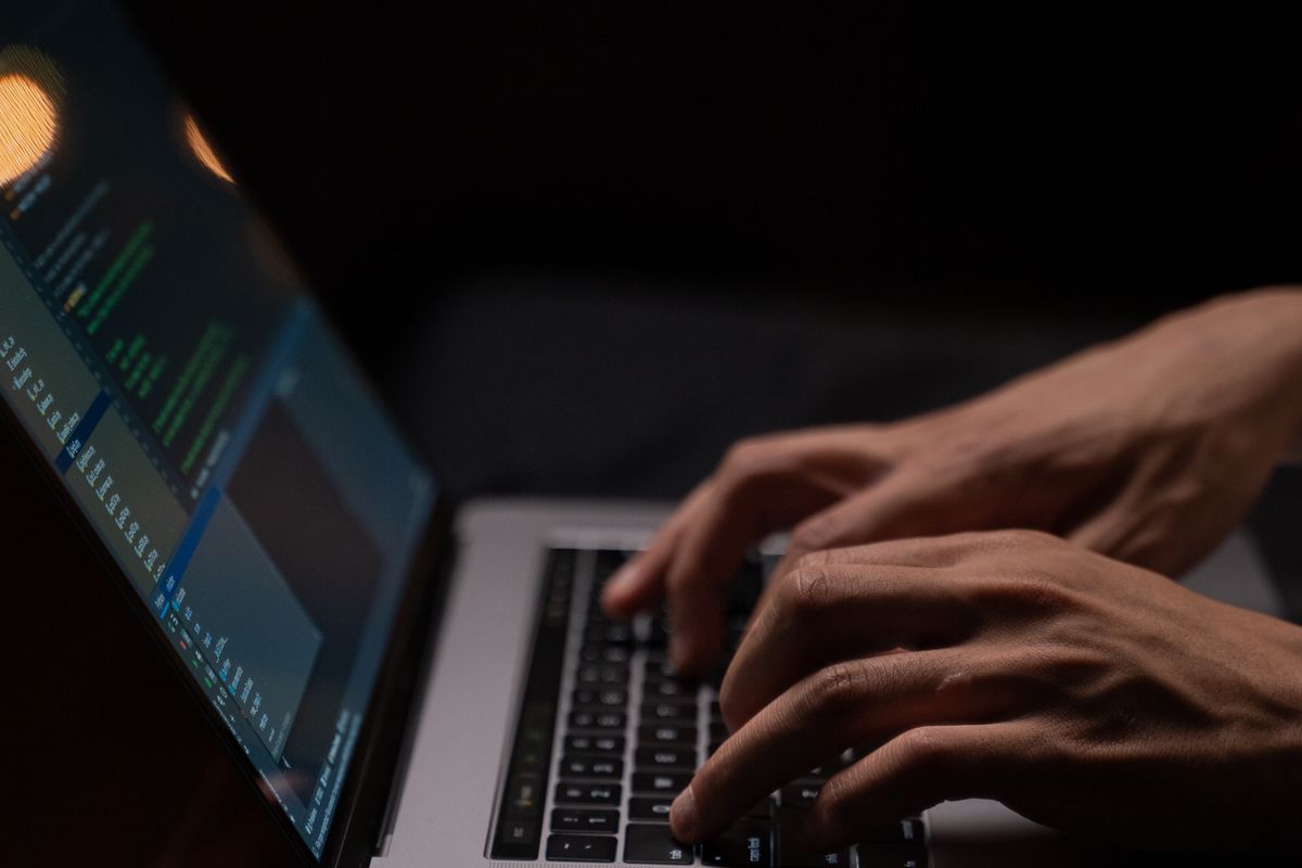Hands typing on a laptop keyboard in a dark room, code glowing on the tilted screen