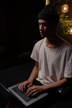 A young man in a beanie and t-shirt working on his laptop in a dimly lit room with soft lighting.