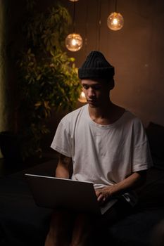 一名戴着无檐帽的年轻人正专注于用笔记本电脑工作，周围挂着吊灯