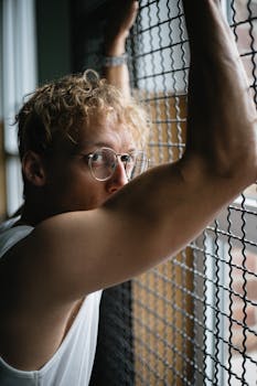 A young man in glasses looking contemplatively out of a window.